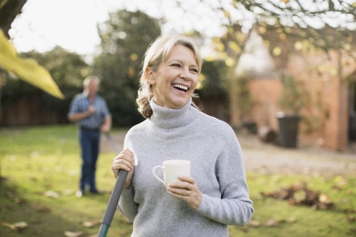 happy mature woman drinking coffee and raking autumn leaves in backyard
