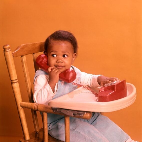 1970s AFRICAN AMERICAN BOY PLAYING WITH TOY TELEPHONE SITTING IN HIGHCHAIR