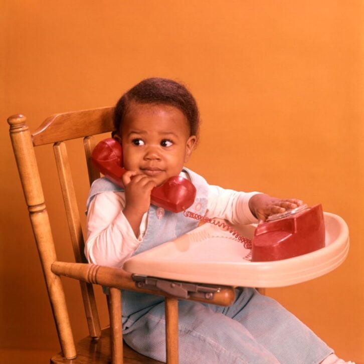 1970s AFRICAN AMERICAN BOY PLAYING WITH TOY TELEPHONE SITTING IN HIGHCHAIR