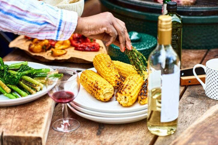Char grilled corn with green vegetables and wine on table, hand seasoning food with herbs