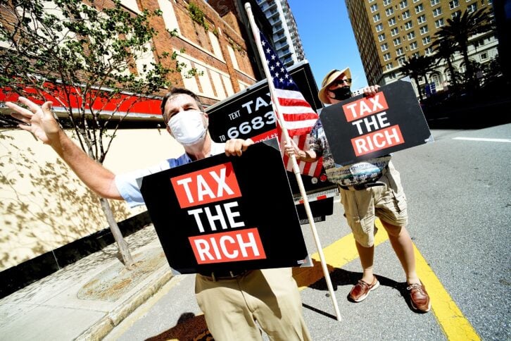 Two men standing outside a senator's office in Florida holding "Tax the Rich" signs.
