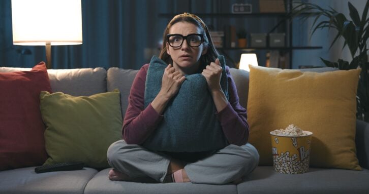 Woman sitting on a couch, watching a horror movie at home and clutching a pillow