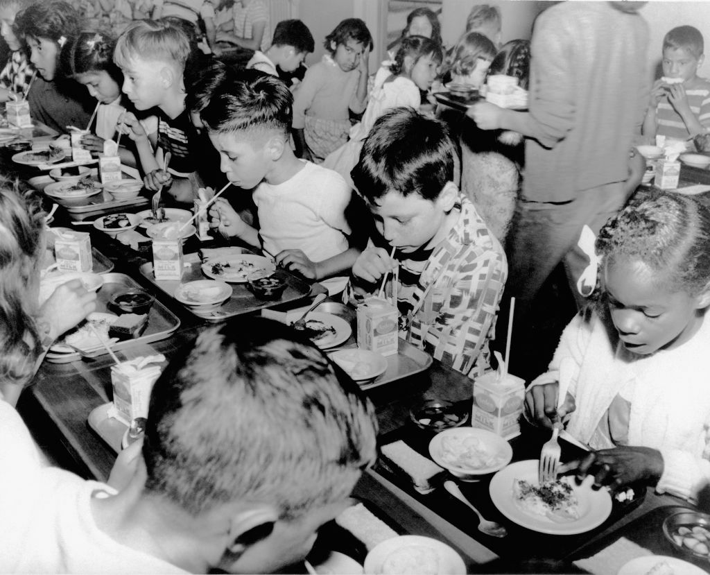 Table full of young children in elementary school eating their lunch off of trays as part of a government funded school lunch program.