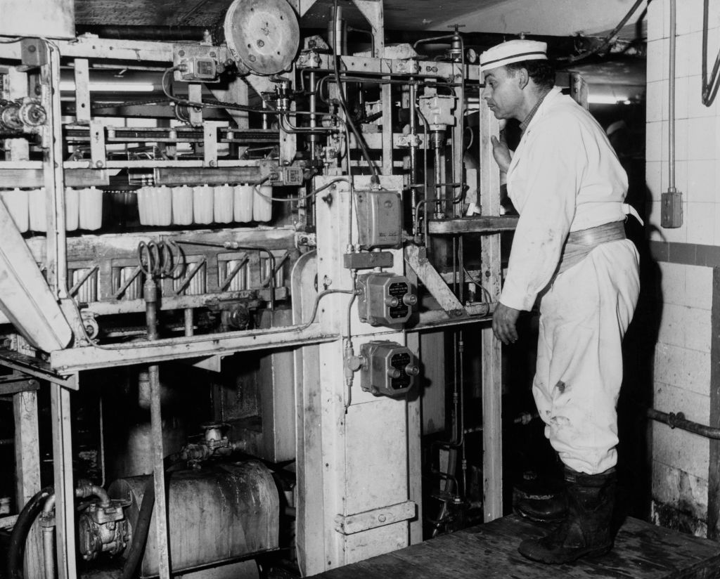 A man wearing coveralls and a cap operates an ice cream-making machine at a factory, United Kingdom, 1962.