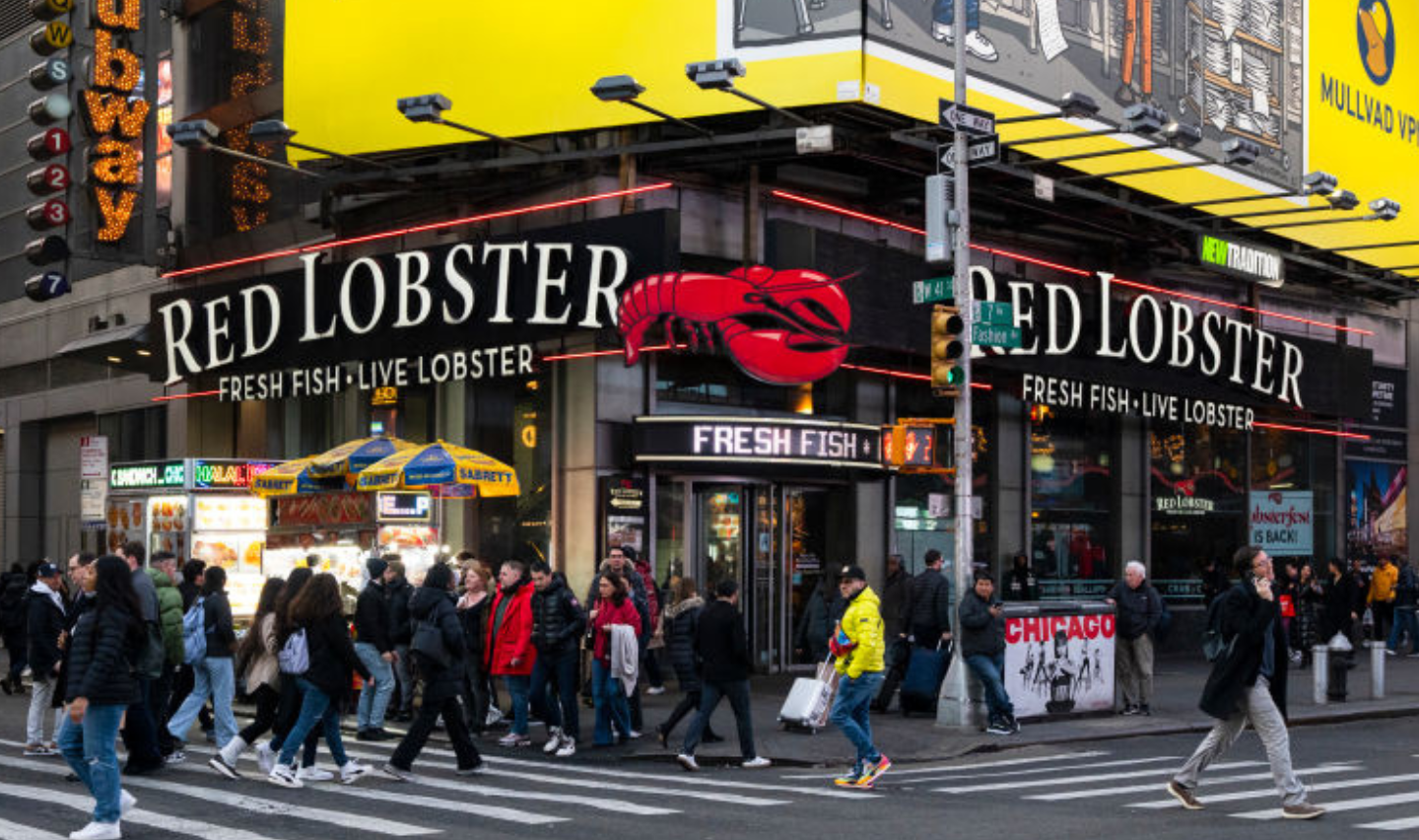 People cross a busy intersection in front of a Red Lobster restaurant with large signs reading "RED LOBSTER" and "FRESH FISH • LIVE LOBSTER" in a lively city setting, likely Times Square, New York.