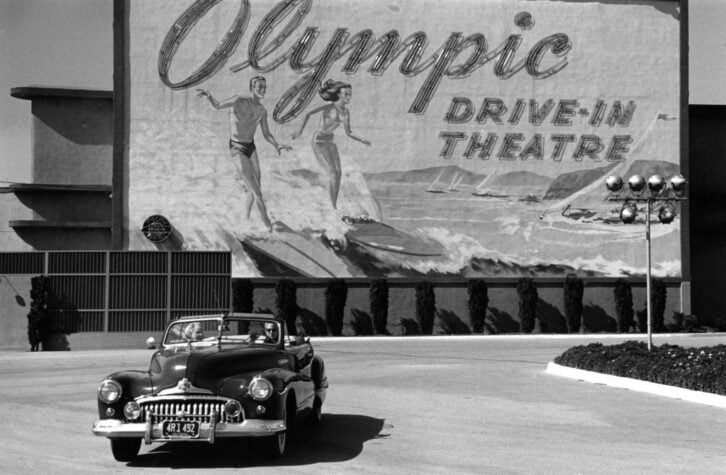 A couple in an open-top car driving past a sign advertising Hollywood's Olympic drive-in theatre in Los Angeles, California. Original Publication: Picture Post - 5298 - Two Men In Hollywood - pub. 1951