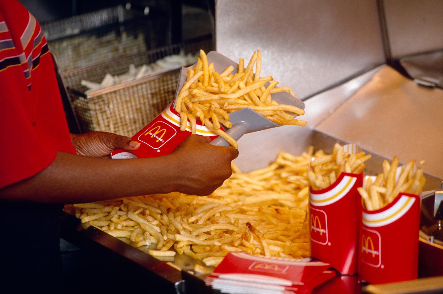 A McDonald's employee uses a scoop to fill red and yellow McDonald's fry containers with hot, golden French fries from a large pile in a fast-food kitchen.