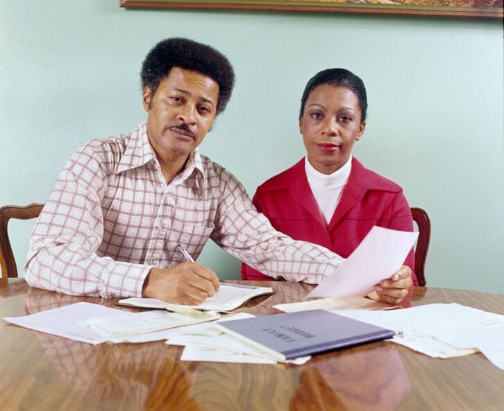 1970s MIDDLE AGED AFRICAN AMERICAN MAN WOMAN COUPLE SEATED TABLE GOING OVER HOUSEHOLD ACCOUNTS LOOKING AT CAMERA