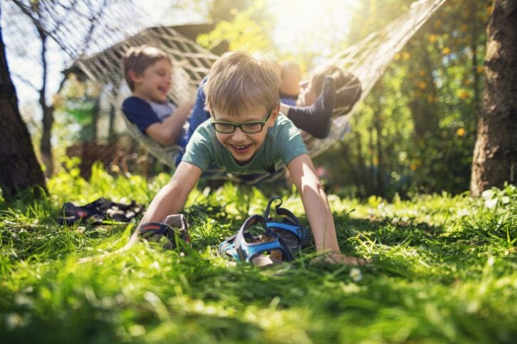 Kids playing in back yard garden on summer day. One of the boys has partially fallen out of the hammock and is laughing