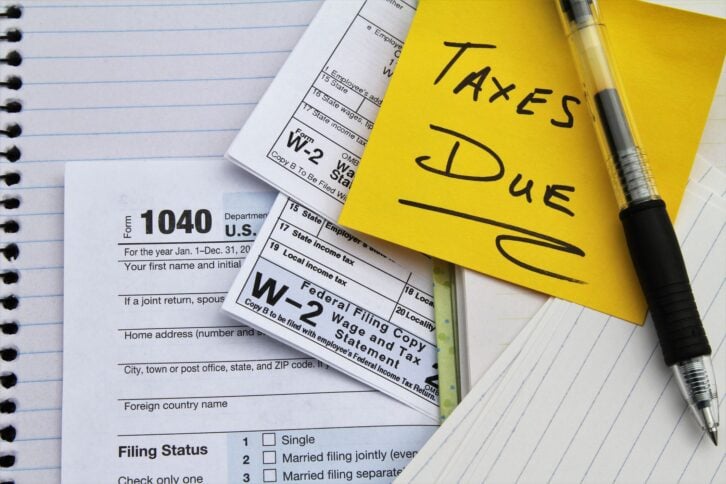 IRS 1040 Tax return form, two W-2 Wage and Tax Statement forms, lined notebook paper, and a pen with a yellow Post-It note saying 'Taxes Due'