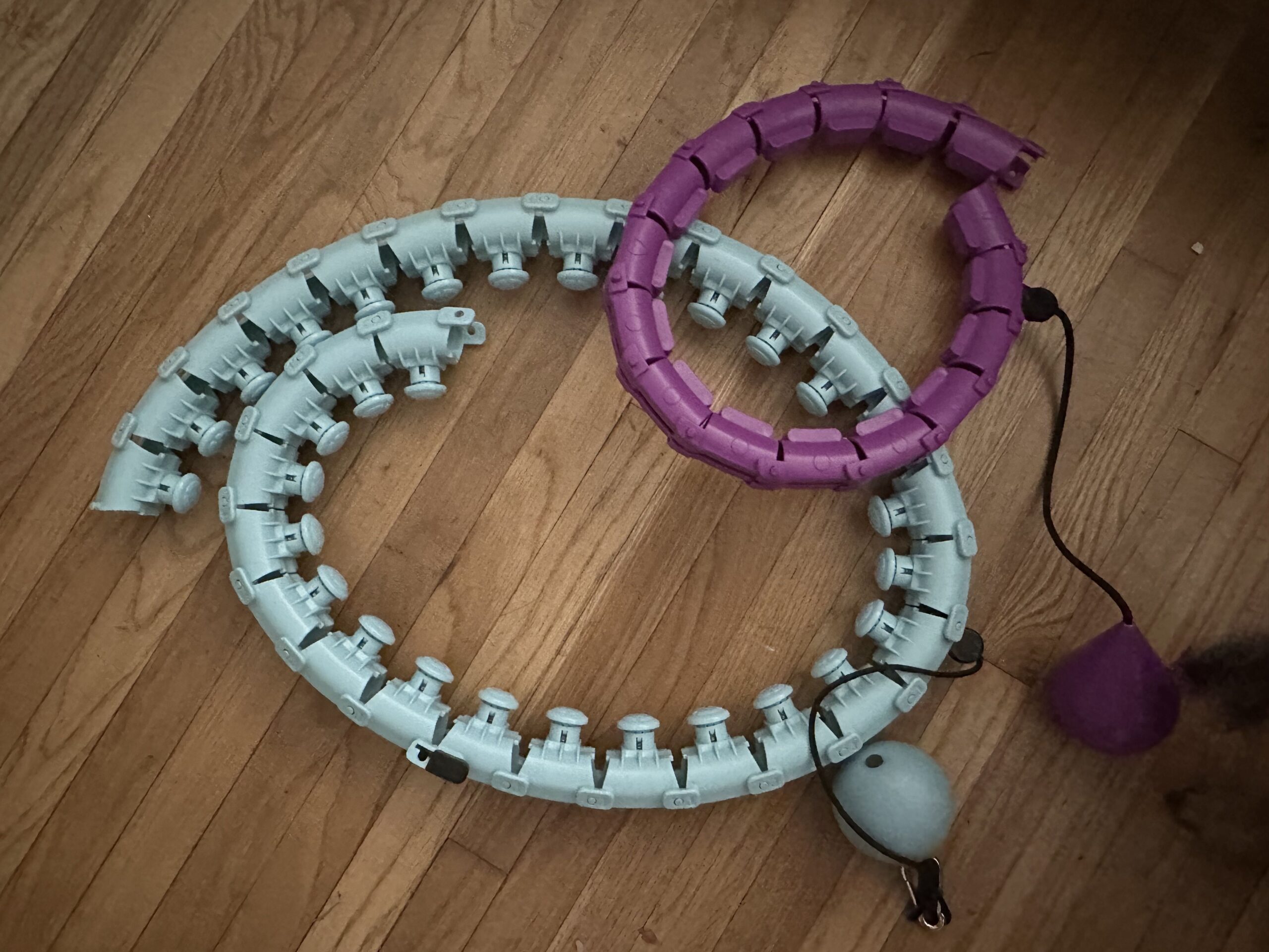 Two adjustable weighted hula hoops, one light blue and one purple, are coiled on a wooden floor. Each hoop has a ball attached to a string for added weight and resistance.