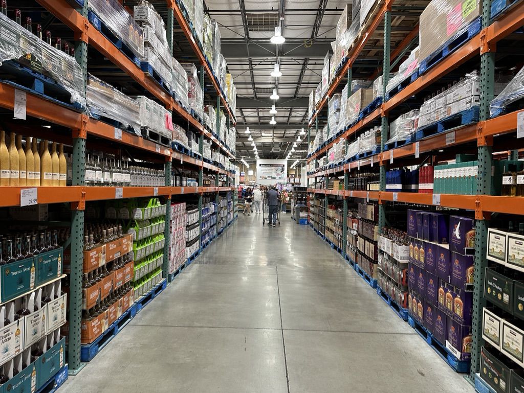 Wide warehouse aisle with tall shelves stocked with boxes, bottles, and various products on both sides, concrete floor, and a few shoppers visible in the distance under bright overhead lighting.