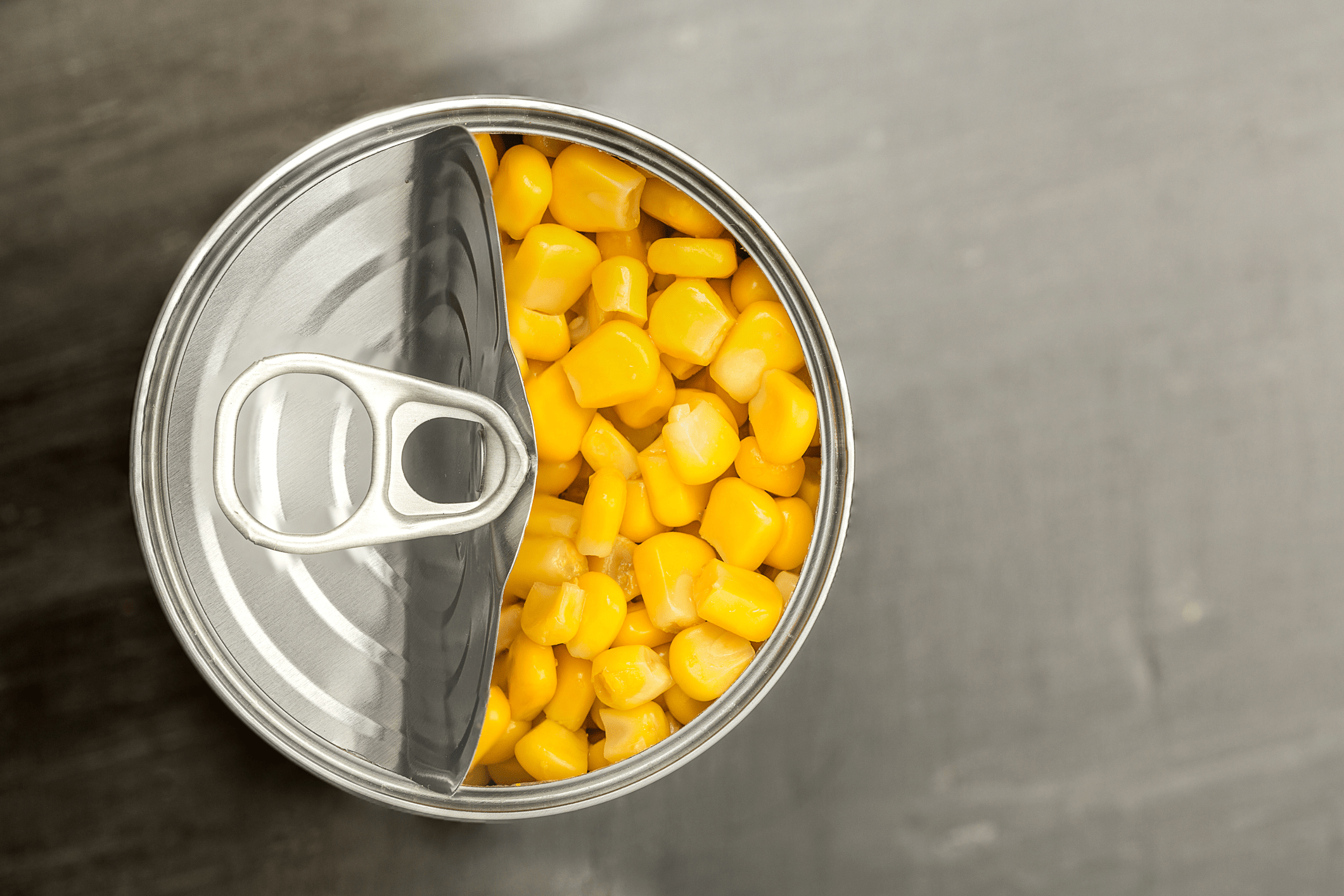 Looking down on an opened can of yellow corn, top pulled halfway, on a wooden table blurred in the background
