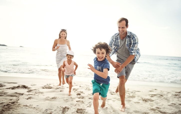 Mother, father, young son, and young daughter running on beach towards the camera, all are smiling and laughing, sand on beach with ocean in the background, light blues and white sky