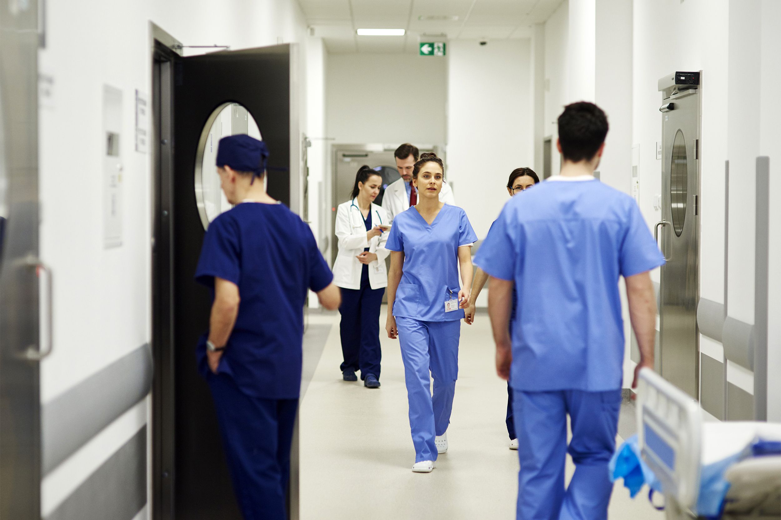 Doctors and nurses in scrubs walk through a bright hospital corridor, some holding medical charts and supplies, with hospital equipment visible along the walls.