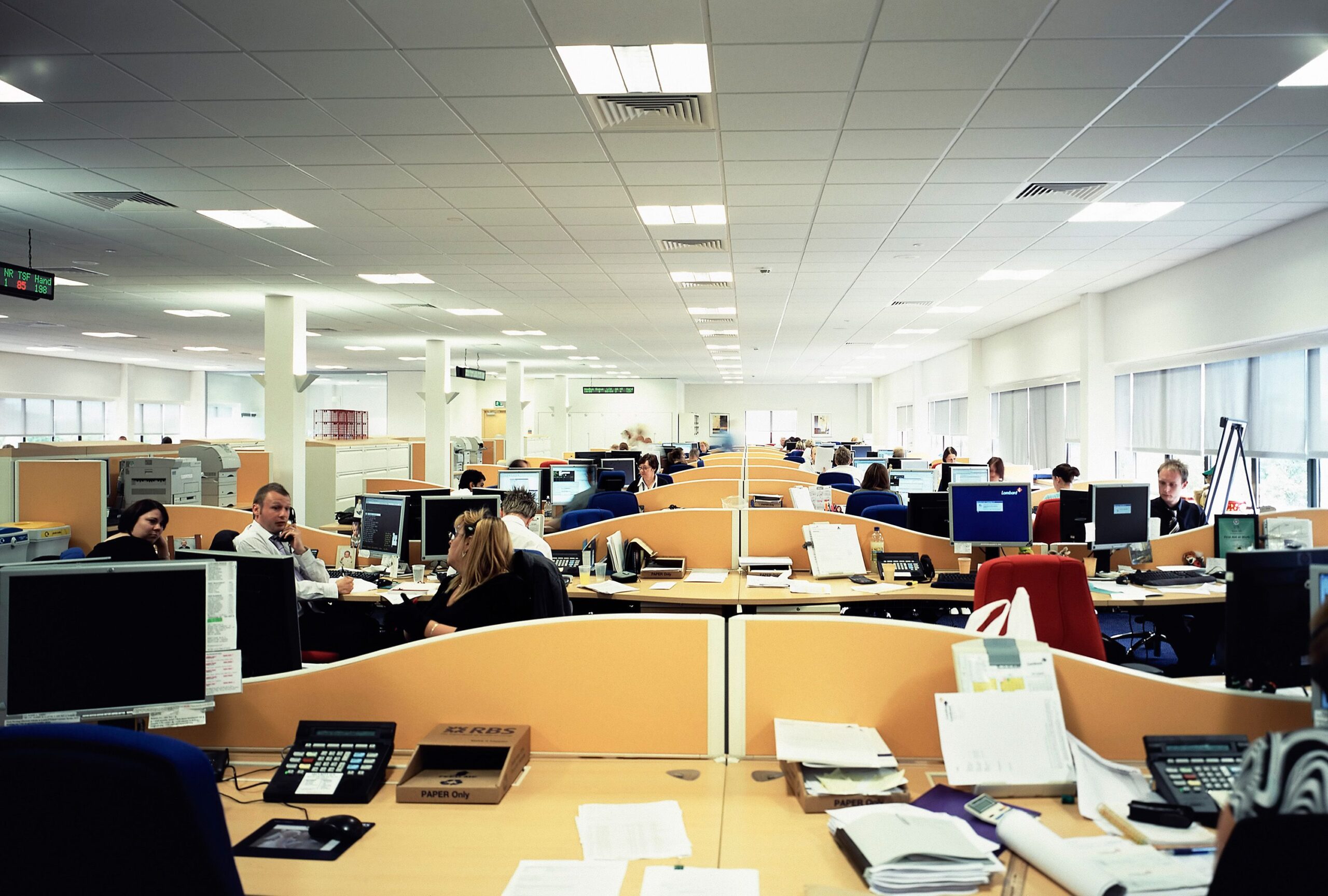 A busy open-plan office with rows of desks and computers, several people working or talking, paperwork and office supplies scattered on desks, and large windows on one side letting in natural light.