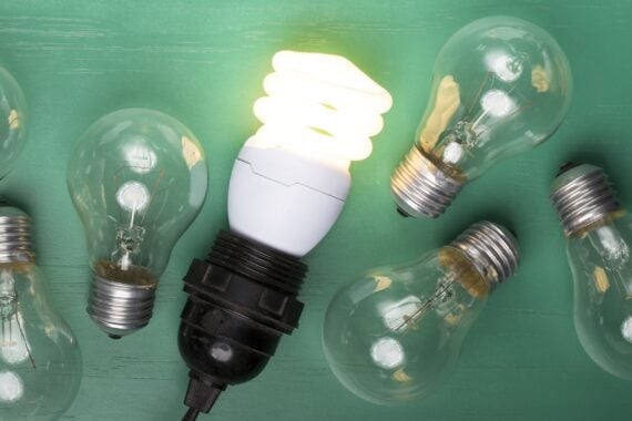 One glowing, energy efficient lightbulb laying next to six older version incandescent lightbulbs, on a dark green wooden table background