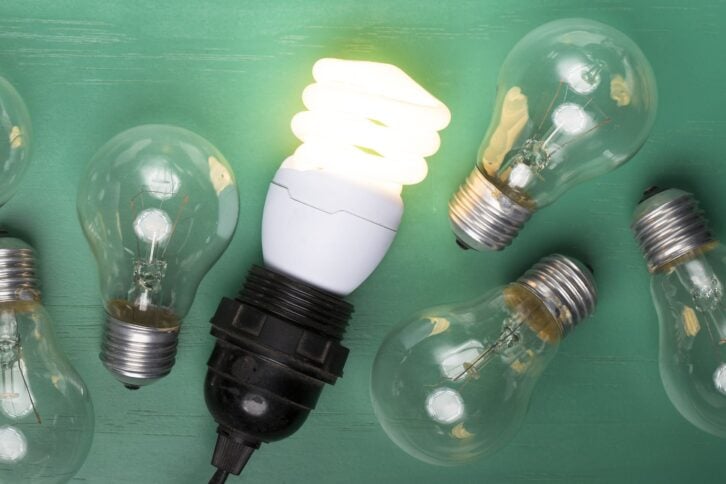 One glowing, energy efficient lightbulb laying next to six older version incandescent lightbulbs, on a dark green wooden table background