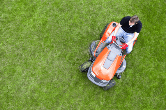 Overhead shot of man on orange riding lawn mower cutting grass, on right side, bright green grass in image