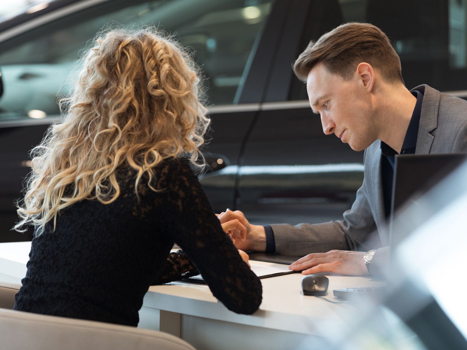 Car salesperson talking with female customer while sitting at desk in showroom