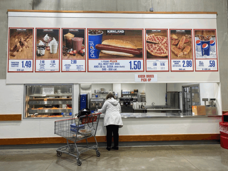 Person getting food at the ordering kiosk in a Costco, large photos of items available with prices on the top of the kiosk, clean concrete floor
