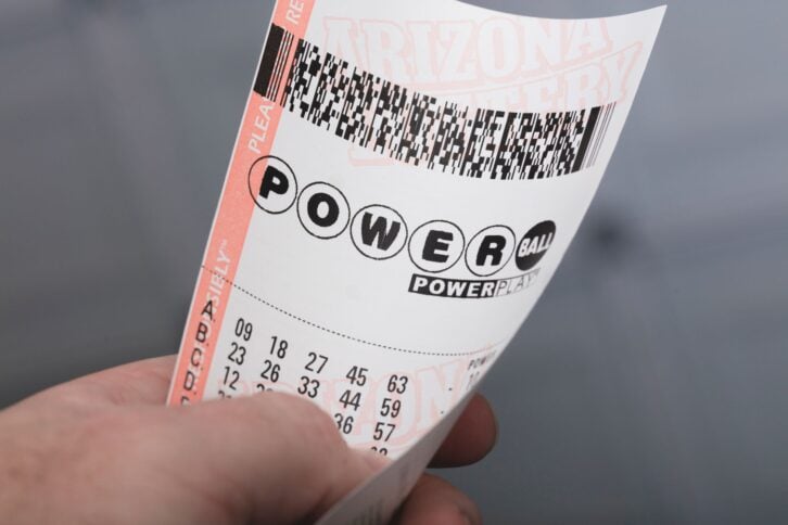Powerball ticket with five sets of numbers, being held by a male left hand, on a blurred grey background