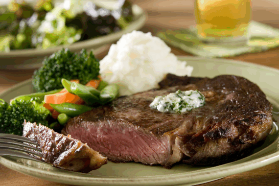 Prime rib-eye steak with mashed potatoes on a beige ceramic plate, one piece of steak on a fork placed on the left, with a blurred background of a salad on a beige ceramic plate and a glass of beer