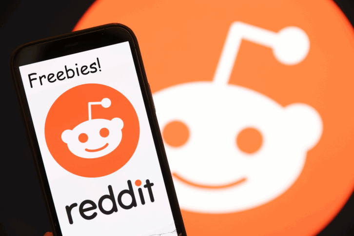 The Reddit logo is displayed on the screen of an iPhone with 'Freebies' above it, to the left side, against a blurred background of a large reddit logo
