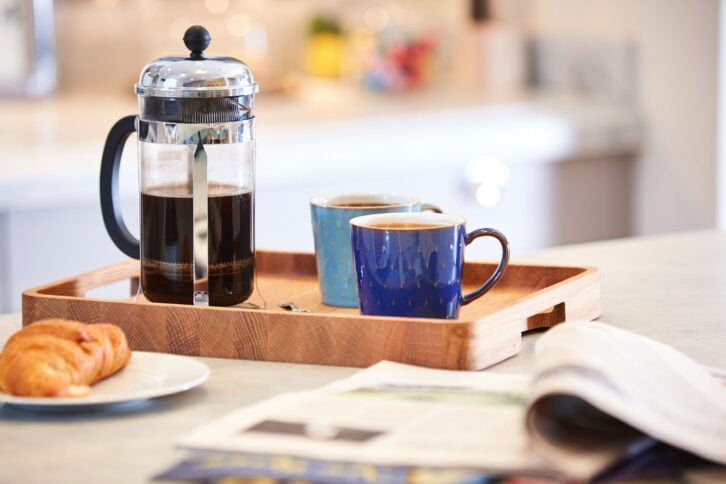 Selective focus of French press carafe filled with dark coffee next to two blue coffee cups, one light blue and the other dark blue, on a wooden serving tray , next to a croissant and newspaper, on a kitchen island with a blurred kitchen countertop in the