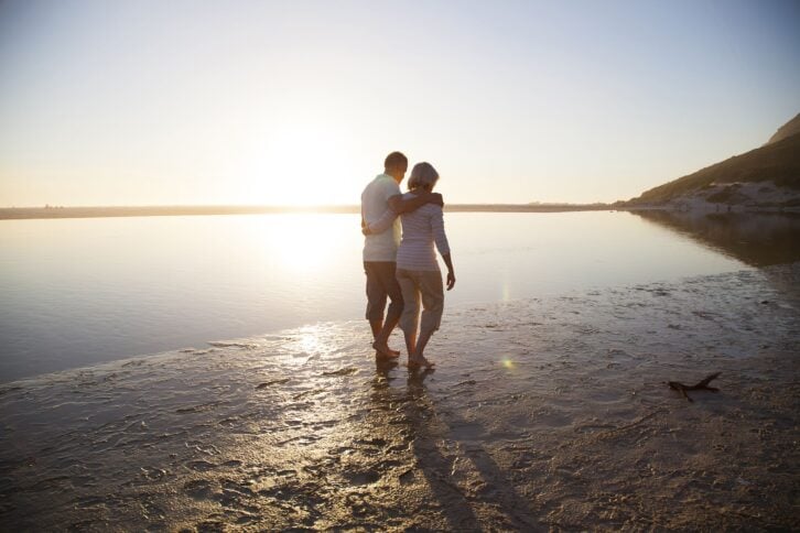 Senior couple walking barefoot on the beach, land on the other side of water, sun is almost setting