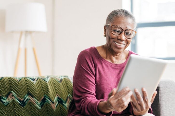 Senior women smiling while holding and looking at a tablet, sitting on a grey couch with a chevron patterned blanket with a lamp and window in the background