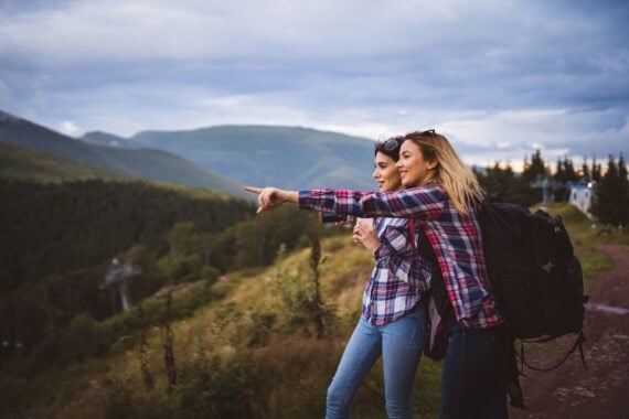 Sides of two woman embraced in a couple in the foreground, one is pointing to the left towards the distance, surrounded by mountains, clouds and dark blue sky in the background