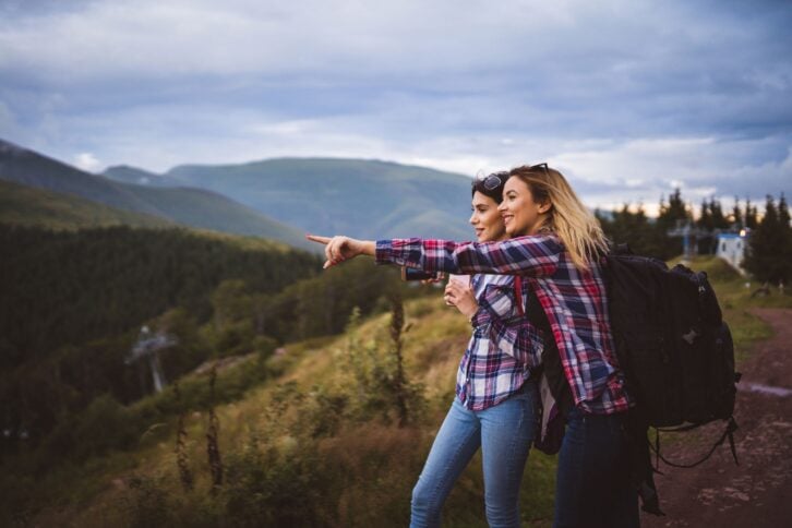 Sides of two woman embraced in a couple in the foreground, one is pointing to the left towards the distance, surrounded by mountains, clouds and dark blue sky in the background