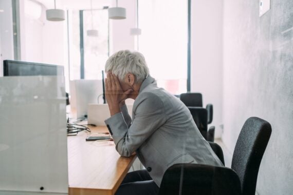 Sideview of a frustrated senior woman sitting at her desk with her hands covering her face, elbows on the desk, in front of a desktop computer, as seen through a glass window, large windows and white walls in the background