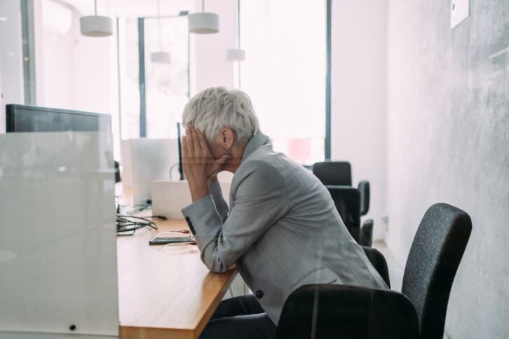 Sideview of a frustrated senior woman sitting at her desk with her hands covering her face, elbows on the desk, in front of a desktop computer, as seen through a glass window, large windows and white walls in the background