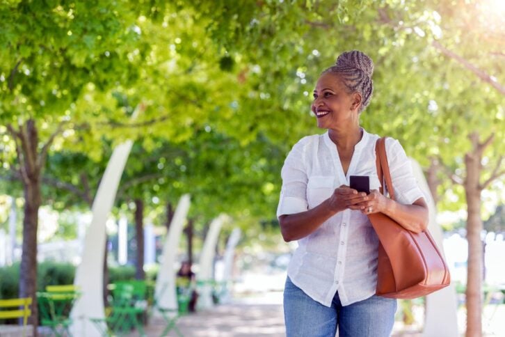 Smiling senior woman holding a smartphone, looking to the left, with a row of trees lining a sidewalk walkway blurred behind her, on a sunny spring day