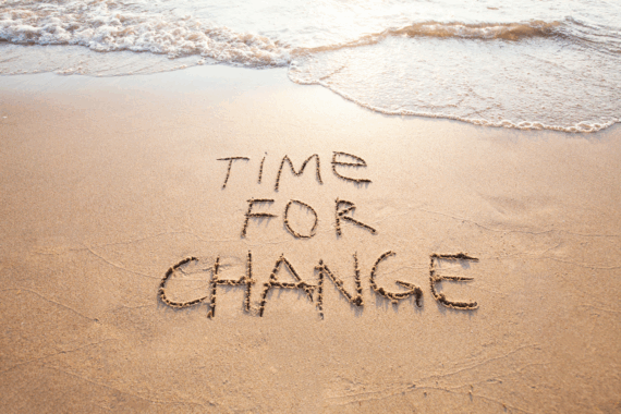 'TIME FOR CHANGE' written in sand in the foreground with ocean waves on the top, sunlight coming through on the top center, during daytime