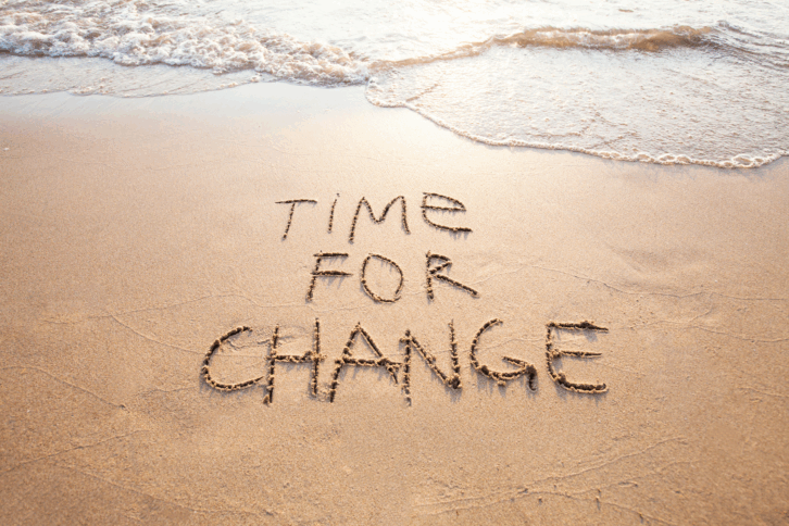 'TIME FOR CHANGE' written in sand in the foreground with ocean waves on the top, sunlight coming through on the top center, during daytime