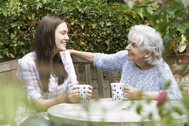 Teenage granddaughter sitting having tea with her grandmother at a table in an outside cafe, with bushes and plants int the background