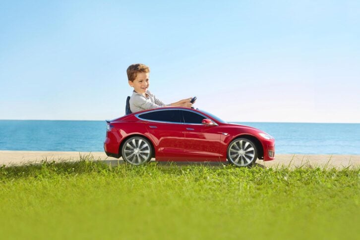 Male child driving a Tesla Model S for Kids by Radio Flyer on a sunny day with the ocean in the background and grass in the foreground