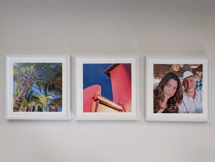 Three framed photos, one of palms, one of red buildings and one of a couple, on a wall in white frames