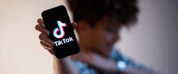 TikTok logo on a smartphone, being held by a male teenager's right hand, selective focus, with a blurred top and midsection of teenager, smiling, against a gray gradient background
