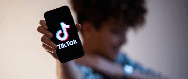 TikTok logo on a smartphone, being held by a male teenager's right hand, selective focus, with a blurred top and midsection of teenager, smiling, against a gray gradient background