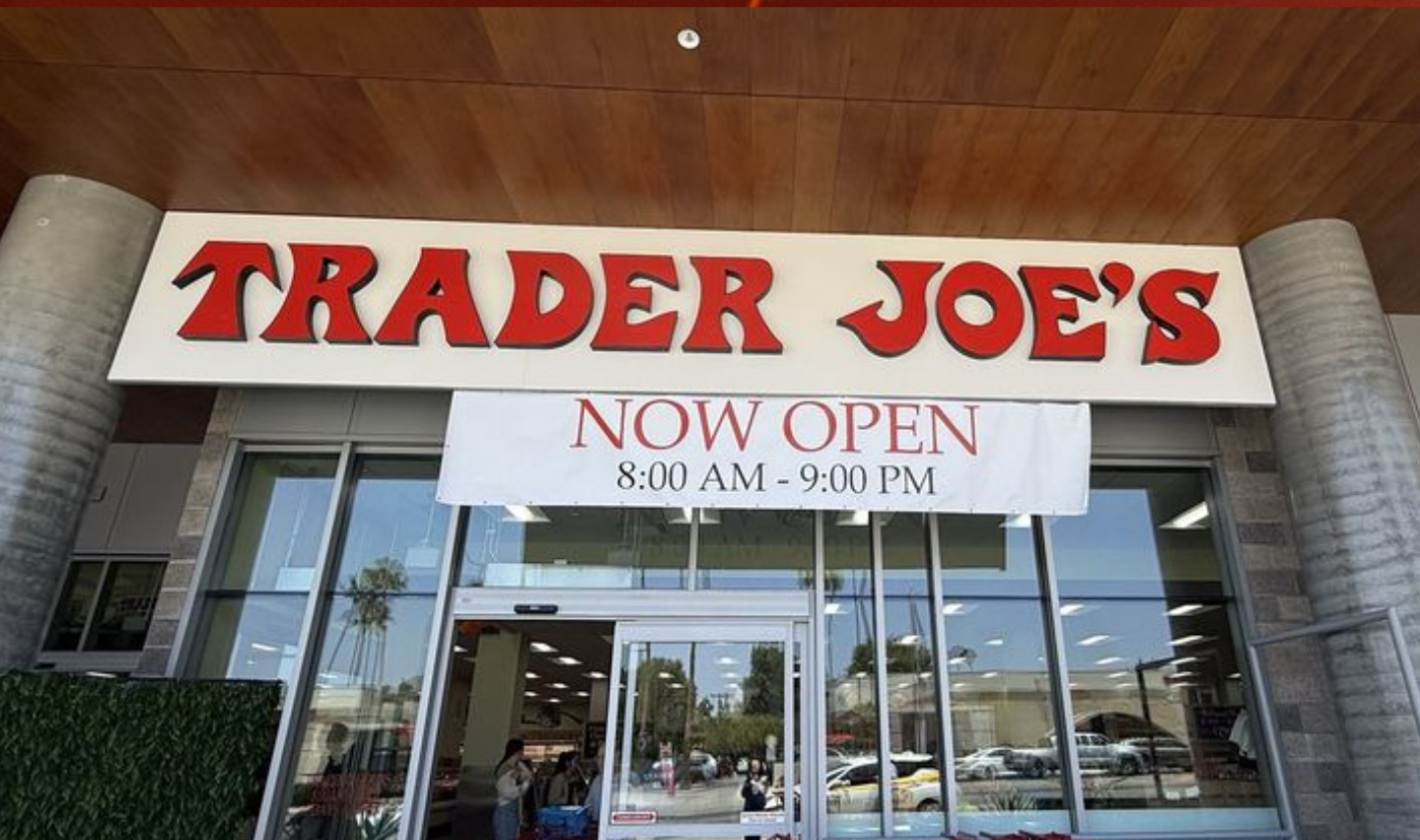 Front entrance of a Trader Joe’s store with a large sign above the doors and a banner below reading “NOW OPEN 8:00 AM - 9:00 PM.” The glass doors show the store interior and people inside.