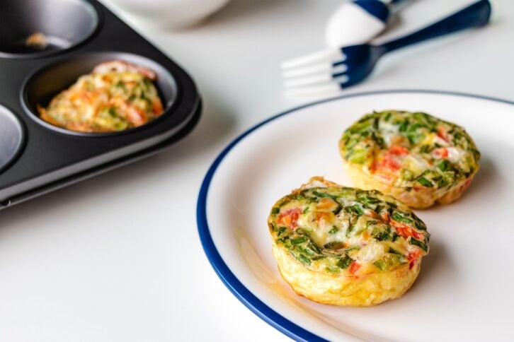 Two breakfast egg muffins with cheese, tomato and green vegetables on a white plate lined with a blue line around the rim with muffins in a muffin tin and utensils blurred in the background