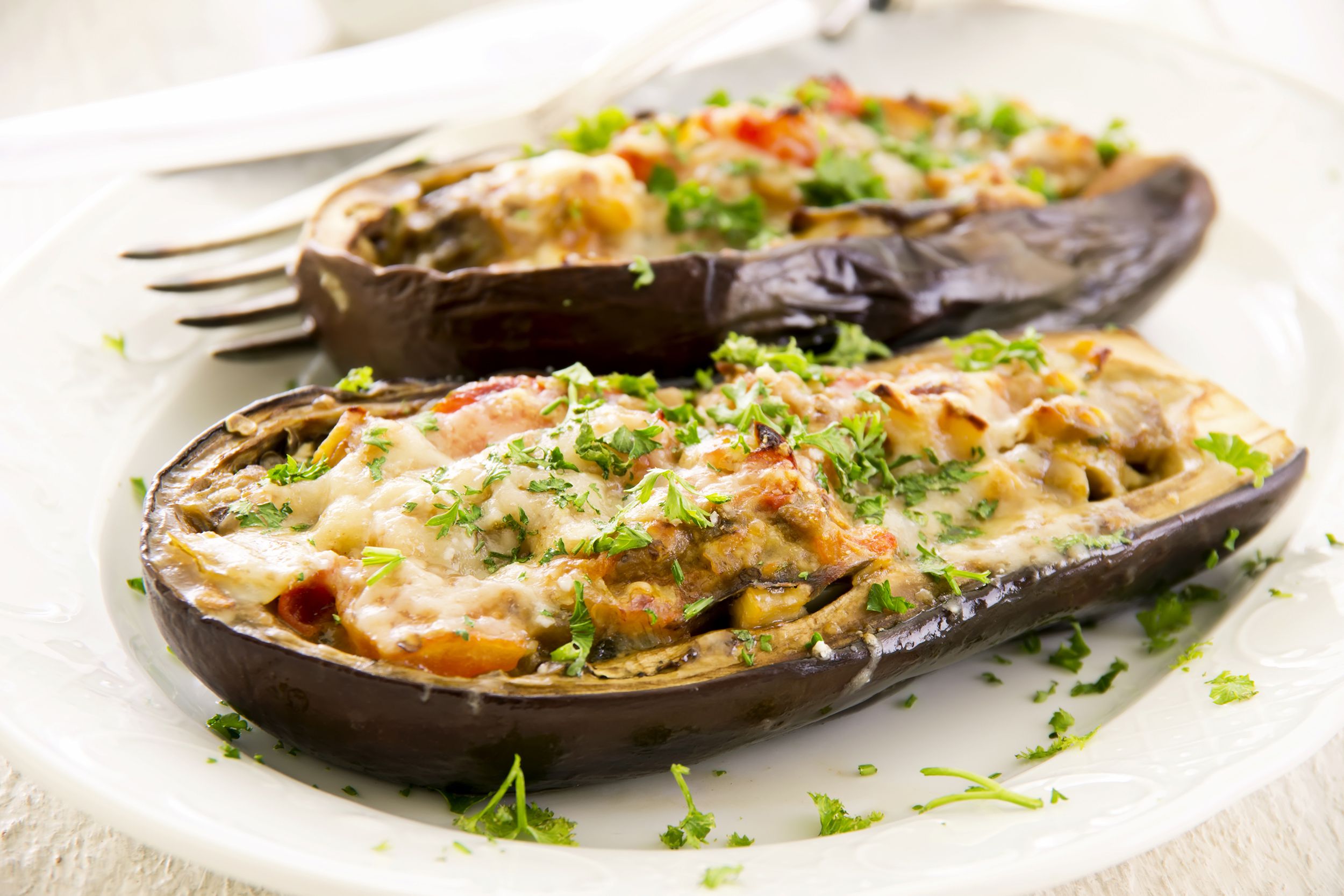 Veggie-stuffed eggplant cut in half on a white porcelain plate, garnished with herbs, with a fork and knife on plate in the top left on a beige wooden table, blurred