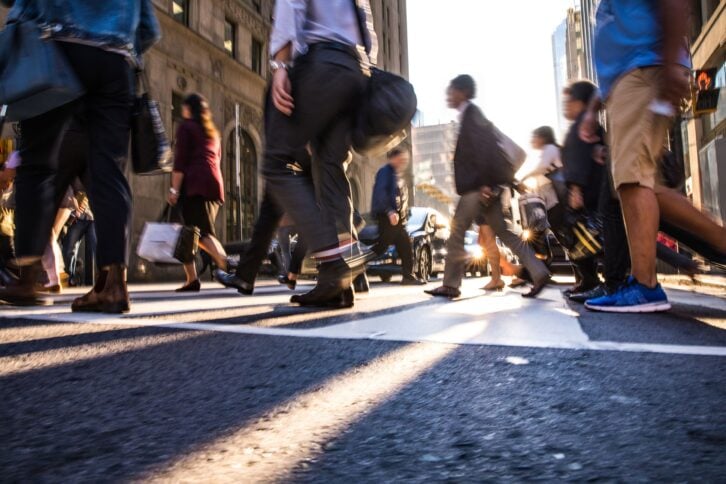 View from below, many people walking along a crosswalk in a city downtown, slight blur, sunlight is coming through buildings in the distance and making lines on the road