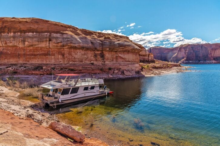White houseboat, on the left, close to the shore of Lake Powell, Arizona, red rock mountains, on a sunny day with a bright blue sky
