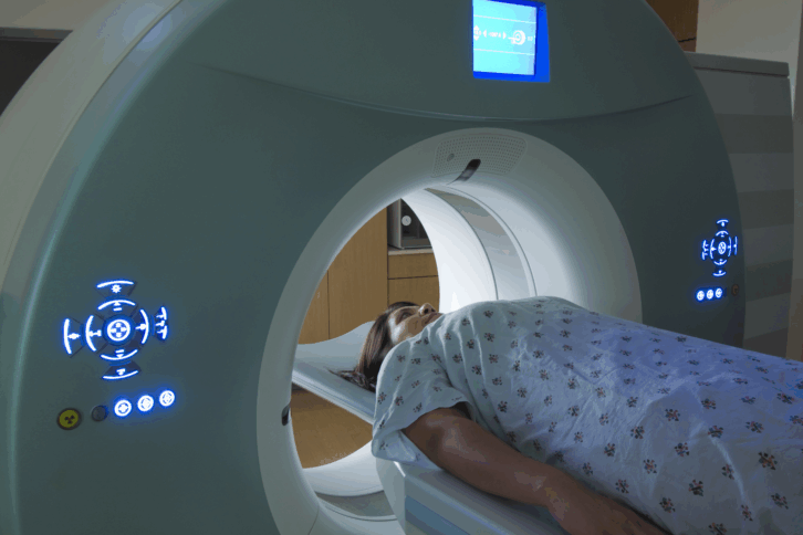 Woman lying down, being scanned by a MRI machine, she is wearing a hospital gown, in a hospital room