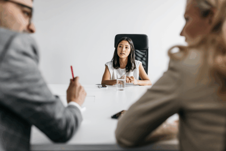 Young woman having a job interview sitting at one end of a white table in the background with a male and female interviewer blurred in the foreground, a clean white wall in the background